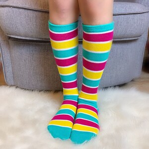 Peut inclure: Une paire de chaussettes montantes &agrave; rayures horizontales turquoise, blanches, jaunes et magenta. Les chaussettes sont port&eacute;es sur les jambes d'une personne, les pieds reposant sur une surface blanche et moelleuse. Les chaussettes ont un bord et un bout turquoise.