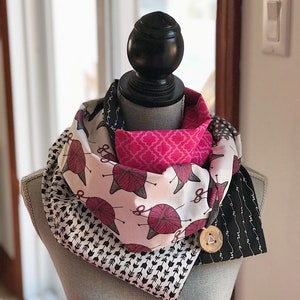 May include: An infinity scarf with multiple patterns, displayed on a mannequin. The scarf includes fabrics with pink yarn balls on white, black arrows, and a black and white design. A wooden button fastens the scarf.