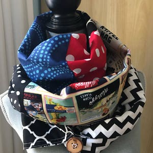 Puede incluir: Bufanda infinita estampada sobre un maniquí. La bufanda presenta secciones de lunares azules, lunares rojos, diseños en blanco y negro y una sección con gráficos vintage de Kellogg's Corn Flakes. Un botón de madera asegura la bufanda.