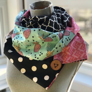 Könnte beinhalten: Ein gemusterter Schal mit Knopfverschluss. Der Schal zeigt Stofflagen mit verschiedenen Designs, darunter schwarze Punkte, Ananas und geometrische Muster. Ein großer Holzknopf sichert den Schal.