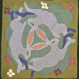 Puede incluir: Un tapiz textil cuadrado que presenta tres liebres de color azul claro dispuestas en un patrón circular sobre un fondo verde salvia. Las liebres tienen detalles cosidos, orejas color melocotón y están rodeadas de pequeñas flores de tela.