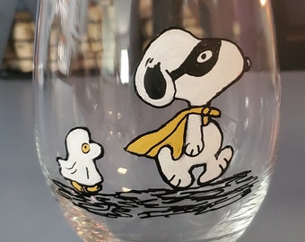 May include: A clear glass wine glass with a hand-painted design of Snoopy wearing a yellow cape and a black mask. A small yellow bird is painted on the glass next to Snoopy.