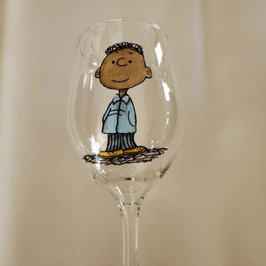 May include: A clear glass wine glass with a hand-painted cartoon character of a young boy with brown skin, black hair, and a blue shirt. The boy is wearing yellow shoes and is standing on a black line.