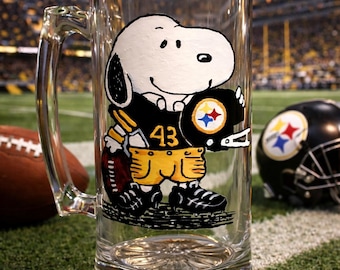 May include: A clear glass beer mug featuring a hand-painted illustration of a white cartoon dog wearing a black and yellow football uniform with the number 43, holding a black helmet with a multi-colored star logo on the side and a football.