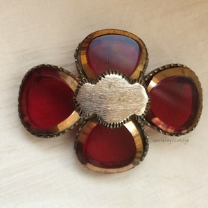 May include: A vintage brooch in a four-petal flower design. The brooch features four red, oval-shaped stones set in a gold-toned metal frame. A silver-toned metal plaque is in the center. The brooch is a unique piece of jewelry.