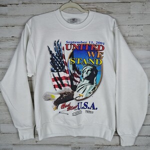 May include: A white long-sleeve sweatshirt featuring a graphic print with the text September 11, 2001 UNITED WE STAND. The design includes an American flag, a bald eagle in flight, and the Statue of Liberty, with the text God Bless the U.S.A.