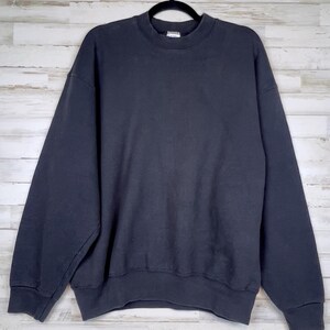 Könnte beinhalten: Ein dunkelmarinefarbenes Sweatshirt mit Rundhalsausschnitt und langen Ärmeln, das an einem schwarzen Kleiderbügel hängt. Der Hintergrund ist eine helle Wand mit Holzpaneelen. Der Stoff wirkt weich und bequem.
