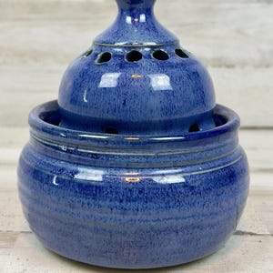 May include: A round, cobalt blue ceramic pot with a matching domed lid. The lid features a series of small, circular ventilation holes around its circumference and a small knob at the top. The glaze has a mottled, speckled texture throughout.