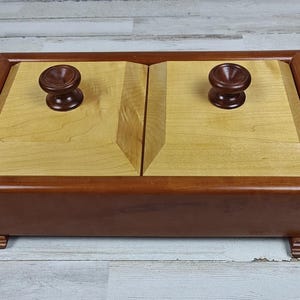 May include: A rectangular wooden box with a light-colored top and dark brown trim. The top is divided into two sections, each with a round, dark brown knob. The box sits on small, square feet.
