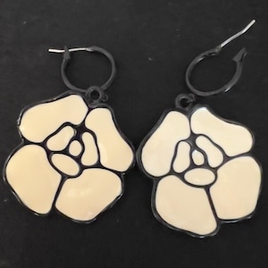 May include: A pair of cream-colored flower-shaped earrings with black outlines and black hoop closures. The earrings have a glossy finish and a floral design, perfect for adding a touch of elegance to any outfit.