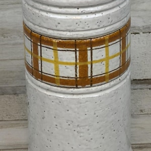 Op de afbeelding: Cilindrische keramische vaas met een gespikkelde witte afwerking. De vaas heeft een band van bruin en geel geruit patroon in het midden. De boven- en onderkant hebben een glad, afgerond ontwerp.