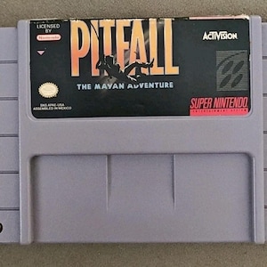 May include: A light purple Super Nintendo video game cartridge for the game Pitfall: The Mayan Adventure. The label features the game title in large yellow and orange letters, the Activision logo, and the Super Nintendo Entertainment System logo.