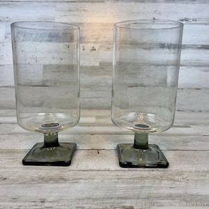 May include: Two clear glass candle holders with a cylindrical shape and a square base. The candle holders have a gray stem and base. The background is a light-colored wooden surface.