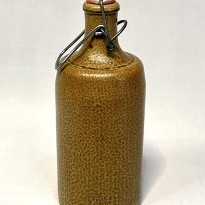 May include: A brown stoneware bottle with a textured surface and a swing-top closure featuring a white ceramic stopper and a metal wire bail. The lower portion of the bottle is embossed with the text 0,7l and MIK-M.