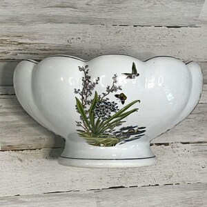 May include: A white ceramic bowl with a scalloped rim and a pedestal base. The exterior features a colorful botanical illustration of green leaves, white flowers, and a small brown butterfly, with a dark bird flying above the floral arrangement.