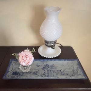 May include: A white hobnail-style lamp with a scalloped top and base sits on a dark wooden surface. A rectangular blue and white decorative tray with a landscape design and a pink flower in a glass vase are also present.