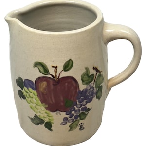 May include: Cream-colored ceramic pitcher with a handle and spout. The pitcher is decorated with a painted design of a red apple, green and purple grapes, and green leaves. The pitcher is suitable for serving beverages.