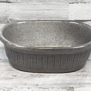 May include: A speckled gray ceramic oval baking dish. The dish has a ribbed design around the exterior and a smooth, rounded rim. The interior is a lighter shade of gray. The dish is designed for baking and serving.