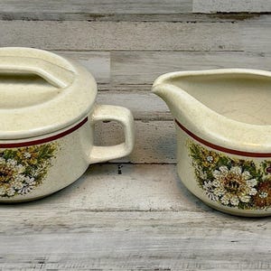 May include: Cream-colored ceramic sugar bowl with lid and creamer set. Both pieces feature a floral design with white, brown, and green flowers, and a dark red band. The sugar bowl has a handle on the lid.