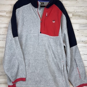 May include: A gray fleece pullover with a quarter-zip closure, featuring a navy blue sherpa collar and shoulder panels. The garment includes a red chest pocket with a zipper, a small logo, and the text Nautica Competition on the left sleeve.