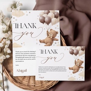 May include: Two baby shower thank you cards with a teddy bear holding balloons. The cards have the words "Thank You" in elegant script. The cards are on a woven basket with white flowers and a brown fabric.
