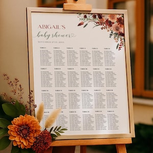 Pode incluir: Um cavalete de madeira exibe um mapa de assentos para um chá de bebê. A placa diz "Abigail's baby shower" com a data. É decorada com arranjos florais em tons de laranja, bordô e verde. Um tecido cor de ferrugem cobre a placa.