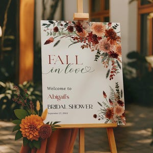 以下が含まれることがあります： 木製のイーゼルには、花柄のアクセントと「FALL in love」の文字が入った看板が飾られています。 看板は、9月27日に予定されているアビゲイルのブライダルシャワーにゲストを歓迎します。 オレンジ色のフラワーアレンジメントと錆色の生地がイーゼルを飾っています。