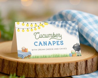 Puede incluir: Un letrero de Canapés de pepino sobre una base de veta de madera clara. El letrero tiene un fondo blanco con las palabras "Canapés de pepino" en verde y azul. El letrero tiene un patrón de cuadros y una guirnalda de luces amarillas.