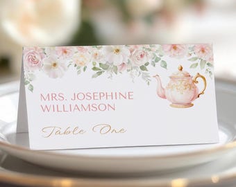 Puede incluir: Una tarjeta de mesa plegada con un diseño floral y una ilustración de tetera rosa. La tarjeta presenta el texto "Mrs. Josephine Williamson" y "Table One" en escritura dorada. El borde superior está adornado con rosas de acuarela.
