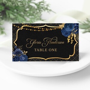 May include: Black and gold wedding place card with a gold frame and blue floral design. The text on the card reads "Gloria Henderson Table One".
