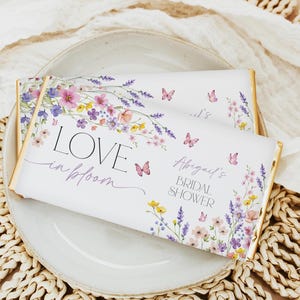 May include: White chocolate bar wrapper with a floral design featuring pink, purple, and yellow flowers and butterflies. The text "LOVE in bloom" and "Abigail's BRIDAL SHOWER" is printed on the wrapper.