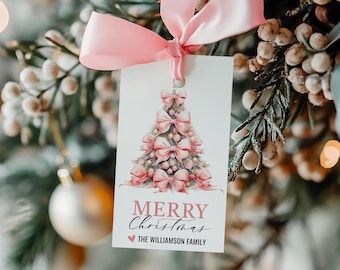 May include: A white gift tag with a pink watercolor illustration of a Christmas tree decorated with pink bows. The text "Merry Christmas" is written in pink lettering with a pink heart and the text "The Williamson Family" below.