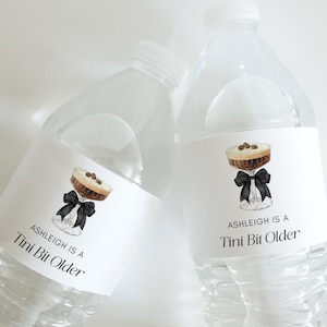 Puede incluir: Dos botellas de agua de plástico transparente con etiquetas blancas. Cada etiqueta presenta una ilustración de acuarela de un cóctel con granos de café y un lazo negro. El texto de la etiqueta dice "Ashleigh is a Tini Bit Older."