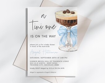 May include: A baby shower invitation with a blue and white bow on a coffee cocktail glass. The invitation reads "a Tini one is on the way" and includes the date, time, and location of the event.