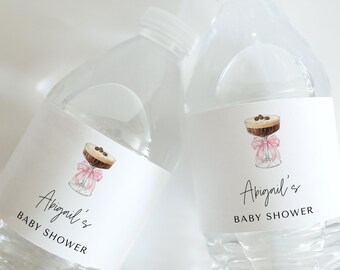 May include: Two clear plastic water bottles with white labels. The labels feature a pink ribbon and a glass of chocolate dessert. The text on the labels reads "Abigail's Baby Shower".