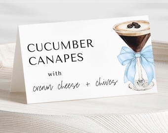May include: White card with black text that reads "Cucumber Canapes with cream cheese + chives". A blue ribbon is tied around a martini glass with a brown drink and coffee beans on top.