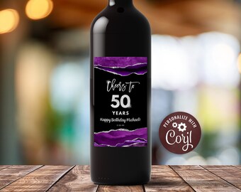 Może przedstawiać: Butelka wina z fioletową i srebrną etykietą, na której widnieje napis "Cheers to 50 Years Happy Birthday Michael". Etykieta ma efekt rozerwanego papieru. Na butelce znajduje się brązowa okrągła naklejka z tekstem "Personalizuj z Cory!"