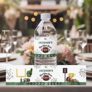 Op de afbeelding: Een doorzichtige plastic waterfles met een wit etiket met een voetbalontwerp en de tekst "Jackson's First Year Down". Het etiket bevat een voetbal, spelschema's en een yardmarker. Extra voetbalthema decoraties zijn zichtbaar.