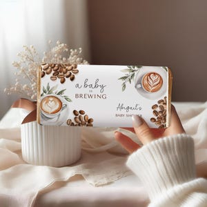 以下が含まれることがあります： 「a baby is BREWING」と「Abigail's BABY SHOWER」の文字が入った白いチョコレートバーの包装紙。コーヒーカップ、コーヒー豆、緑の葉のデザイン。白いセーターを着た人が包装紙を持っています。