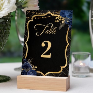 May include: Black and gold table number card with a gold frame and navy blue floral accents. The card says "Table 2" in gold lettering.