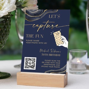 May include: Navy blue and gold birthday party sign with the text "LET'S capture THE FUN". The sign includes a QR code and a graphic of a hand holding a phone. The sign is displayed on a wooden block.