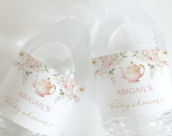 Puede incluir: Dos botellas de agua de plástico transparente con etiquetas. Las etiquetas presentan una ilustración de tetera rosa, detalles florales y el texto "ABIGAIL'S baby shower" en rosa y dorado. Las botellas están llenas de líquido transparente.