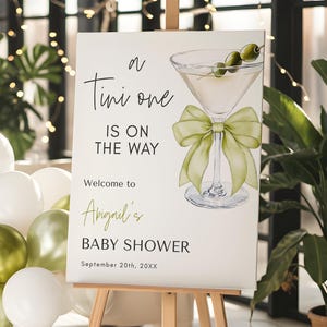 Könnte beinhalten: Ein weißes Schild mit grünen Akzenten, das eine Babyparty für Abigail ankündigt. Das Schild zeigt ein Martini-Glas mit Oliven und einer grünen Schleife. Der Text lautet "A Tini one is on the way. Welcome to Abigail's Baby Shower. September 20th, 20XX".