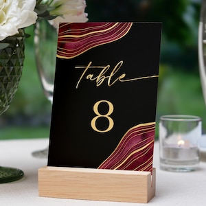 May include: Black table number card with gold lettering and a burgundy and gold agate design. The card reads "Table 8" and is displayed on a wooden stand.