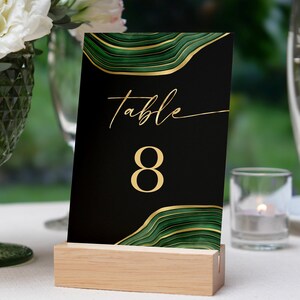 May include: A black table number card with gold script that reads "table" and the number "8". The card has a green and gold abstract design and is held in a light wood block.