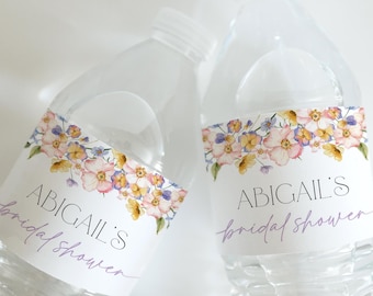 May include: Two clear plastic water bottles with labels featuring a floral design in shades of pink, yellow, and purple. The labels also include the text "Abigail's bridal shower" in a purple script font.
