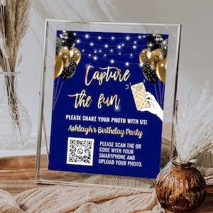 May include: Blue and gold birthday party sign with the text "Capture the fun" and a QR code. The sign also includes a cartoon illustration of a hand holding a smartphone with a QR code on the screen. The text "Please share your photo with us! Ashleigh's Birthday Party" is also included on the sign.