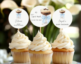 May include: Three cupcakes with white frosting, each topped with a circular topper on a wooden stick. The toppers feature a watercolor illustration of a cocktail with a blue bow and text that reads "Welcome Baby Hutchinson", "A Tiny One Is On The Way", and "Abigail's Baby Shower".
