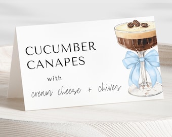 May include: White card with black text that reads "Cucumber Canapes with cream cheese + chives". A blue ribbon is tied around the stem of a glass filled with a brown liquid and topped with whipped cream and coffee beans.