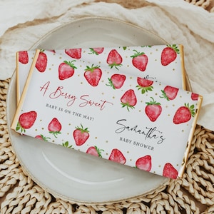 May include: A white chocolate bar wrapper with a gold border. The wrapper features a red and green strawberry pattern with gold glitter. The text "A Berry Sweet Baby is on the way!" and "Samantha's Baby Shower" is printed on the wrapper.